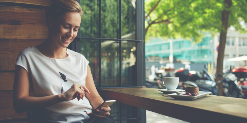 A woman sitting in a cafe with a coffee and food next to her while happily scrolling on her phone.